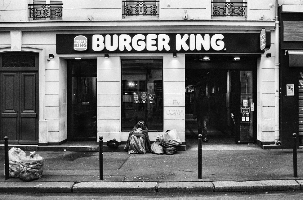 Black and white photo of a Burger King storefront. In front, a person is sitting on the sidewalk surrounded by bags and belongings. The street in front has poles and some trash. The building exterior features ornate wrought iron railings on the windows above, suggesting an unexpected contrast to the fast food scene below.