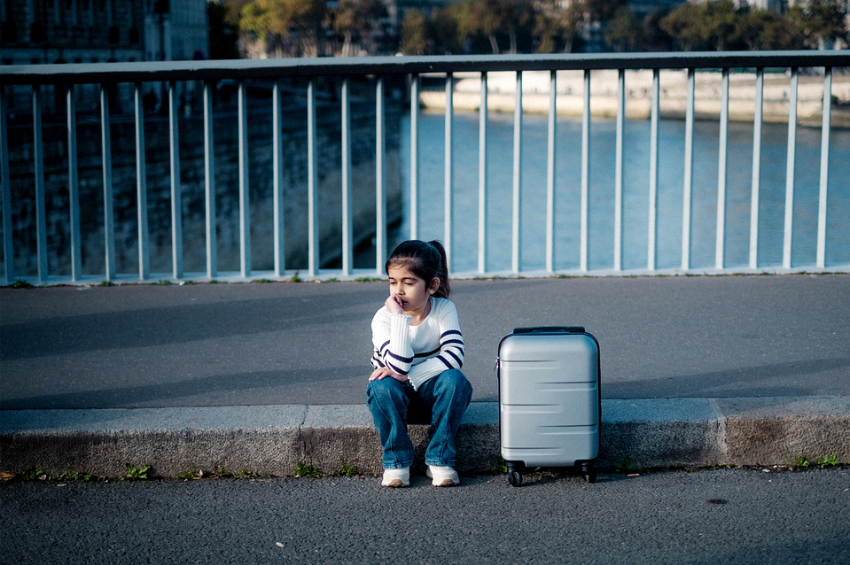 A young child sits on a curb by a river, beside a gray suitcase. They wear a white sweater with black stripes and blue jeans. A metal railing separates the sidewalk from the tranquil water in the background, creating an atmosphere of calm contemplation.