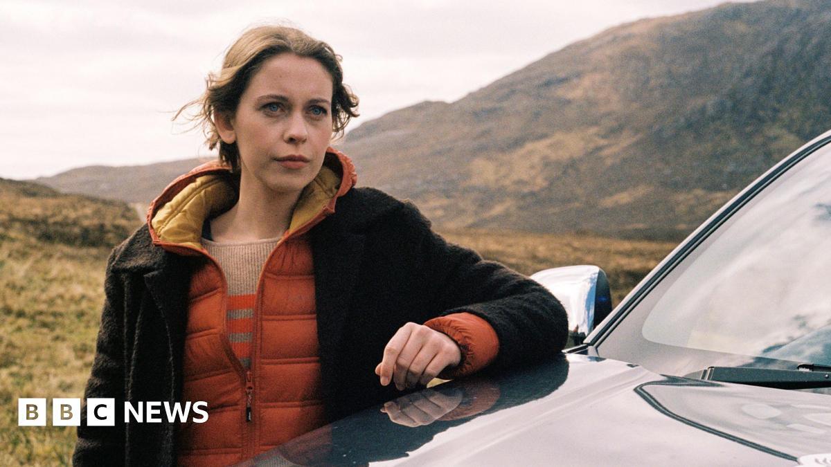 Star Sorcha Groundsell as Kat in An t-Eilean (The Island) - she is outside leaning on a car bonnet, with the hills pictured behind her. She has auburn hair, a pensive look on her face and a brown and orange jacket on.