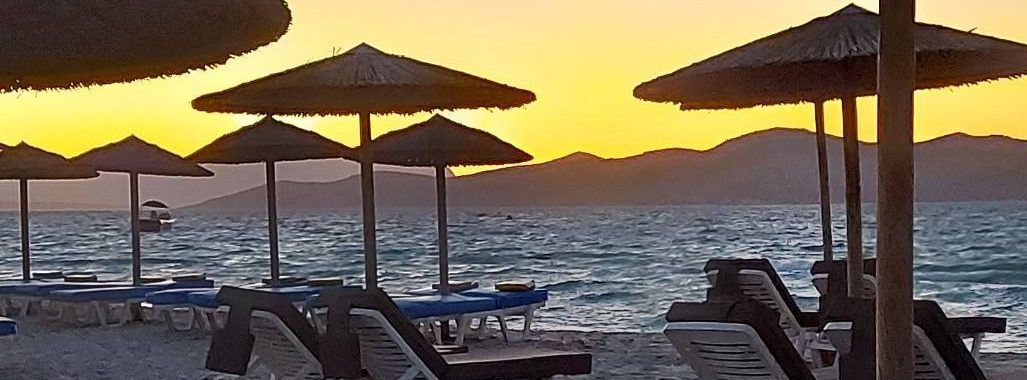 Image: Holly Kos beach, showing sun loungers by the sea, and the setting sun behind distant mountains.