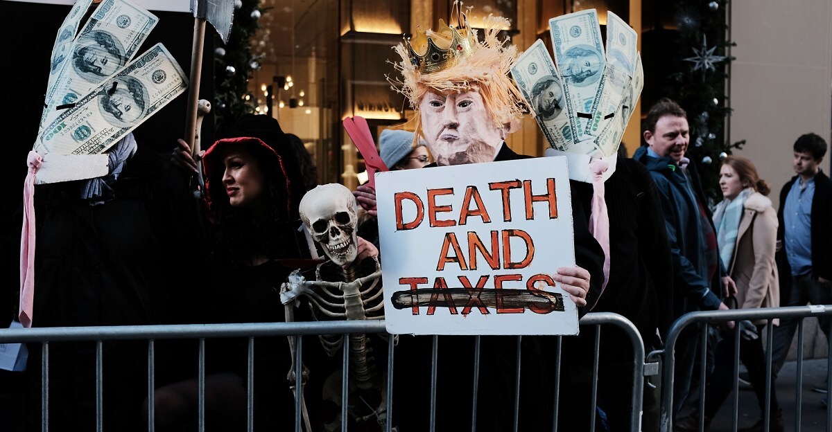 A protestor wearing a Donald Trump paper mâché head stands in front of a barricade and holds a sign that reads, “Death and taxes.”