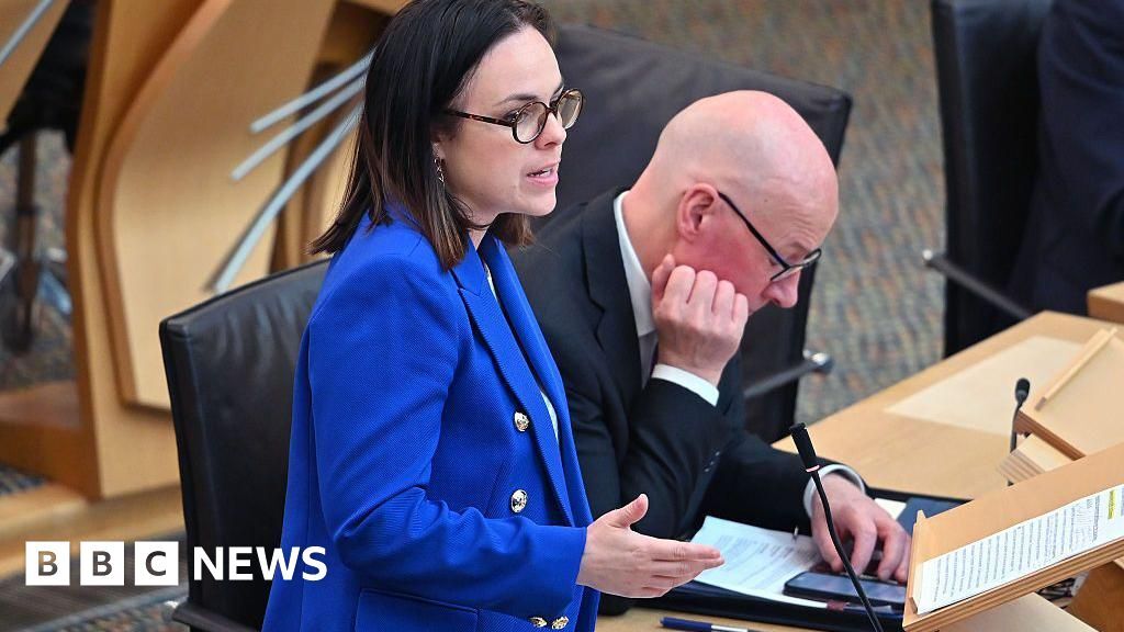 Kate Forbes is speaking in the Scottish Parliament with John Swinney sitting next to her. Forbes has dark shoulder-length hair and is wearing black framed glasses and a blue jacket over a white top. Swinney is looking down at his phone. He is a bald man with glasses and is waring a black jacket over a white shirt