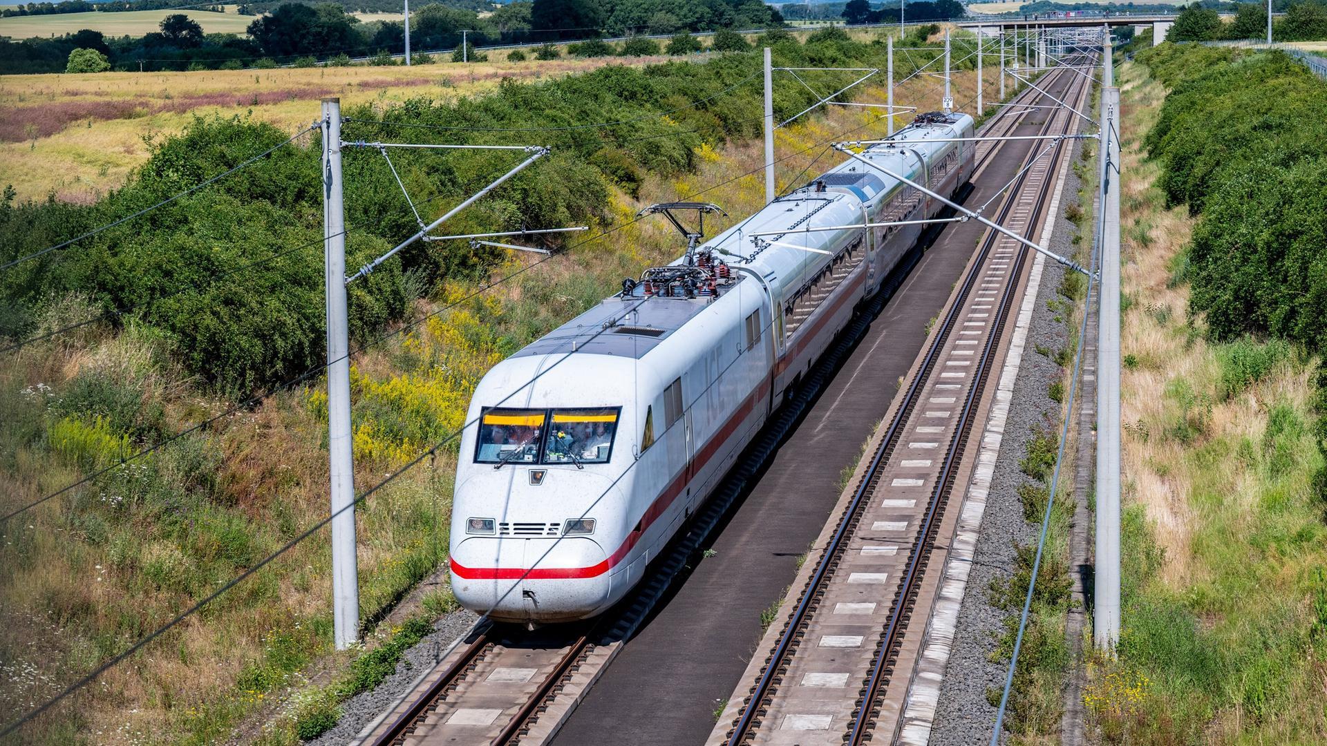 Ein weißer Zug mit rotem Streifen und etwas kantigem Vorderteil fährt auf Schienen durch eine Landschaft.