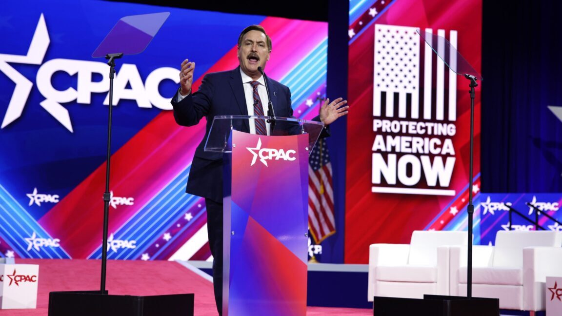 Mike Lindell speaks on stage at the Conservative Political Action Conference, flanked by signs that say "CPAC" and "Protecting American now."
