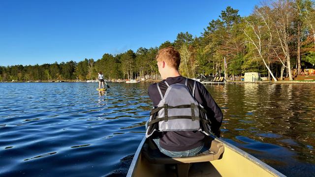 A person in a life vest is canoeing on a calm lake, looking towards another person paddleboarding in the distance. The lake is surrounded by a dense forest under a clear blue sky.