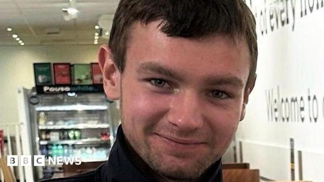 A man wearing a black round-necked top. He has short, brown hair and stubble and he is smiling.