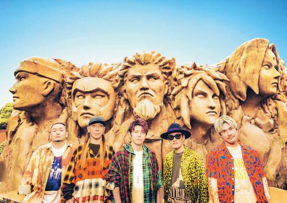 Pictured are the five members of FLOW in colourful clothing in front of the giant Hogake Rock at the Nijigen no Mori theme park.