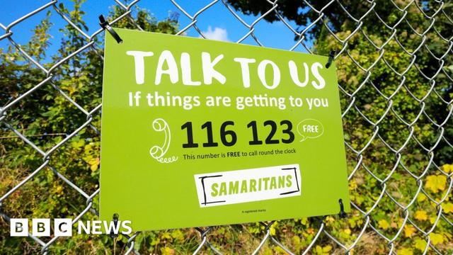 An image shows a Samaritans sign on a railing in front of some bushes. The sign is green with black and white writing and gives their contact number, alongside the words 'talk to us if things are getting to you' - the number listed is 116123 - taken in the UK in 2017.