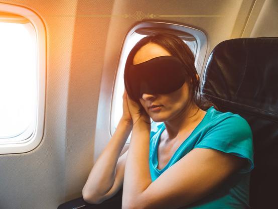 A woman on a plane, wearing a green tshirt and a black eye mask, leaning on the window, sleeping.