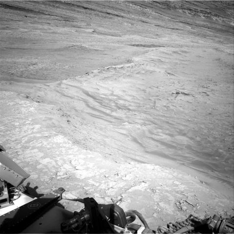A grayscale photo from the Martian surface shows rocky, uneven, light-gray terrain that slopes downward, away from the rover. Parts of the rover are visible in the bottom of the frame, and the downward slope extends from the bottom of the frame toward the top of the frame, with a ridge line tracing the descent.