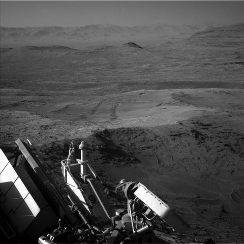 A grayscale photo from the Martian surface shows a landscape of dark gray, slightly uneven, desert-like terrain stretching off into the distance, where a line of peaks that look like sand dunes create a hazy horizon line. Part of the rover is dimly visible in the bottom of the frame, and a pair of wheel tracks, darker than the surrounding ground, extend backward from there, scratched into the soil at the center of the frame.