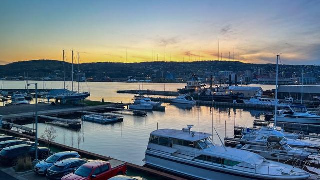 Boats docked at a marina with calm water at sunset, city buildings and a hill in the background, and parked cars in the foreground. The sky is blue and orange, creating a peaceful evening scene.