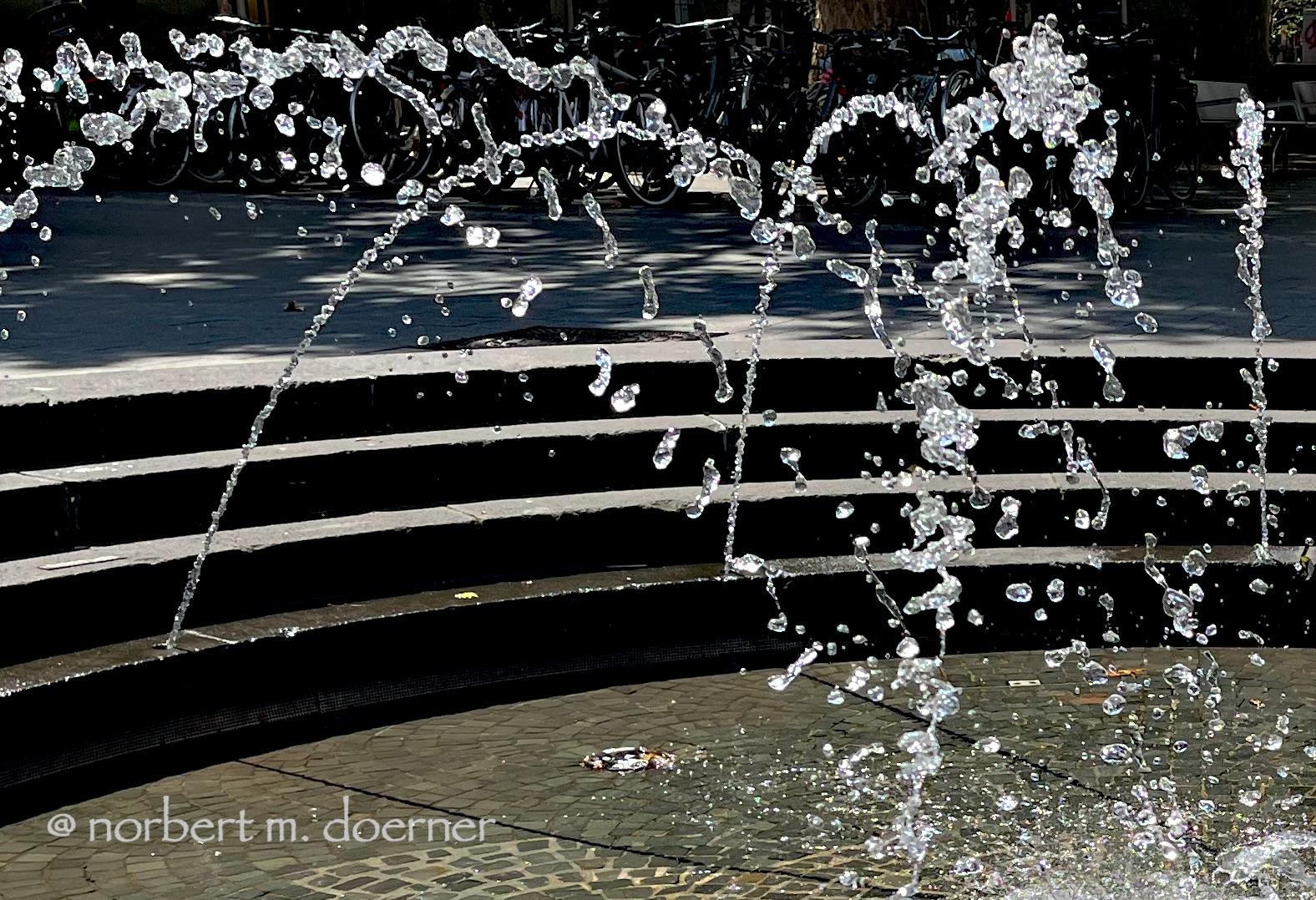 Mehrere Wasserfontänen in einem runden, gemauerten Springbrunnen, die sich in der Mitte treffen, und statt eines Wasserstrahls so aussehen, als wären es einzelne Wasser-Blobbs.