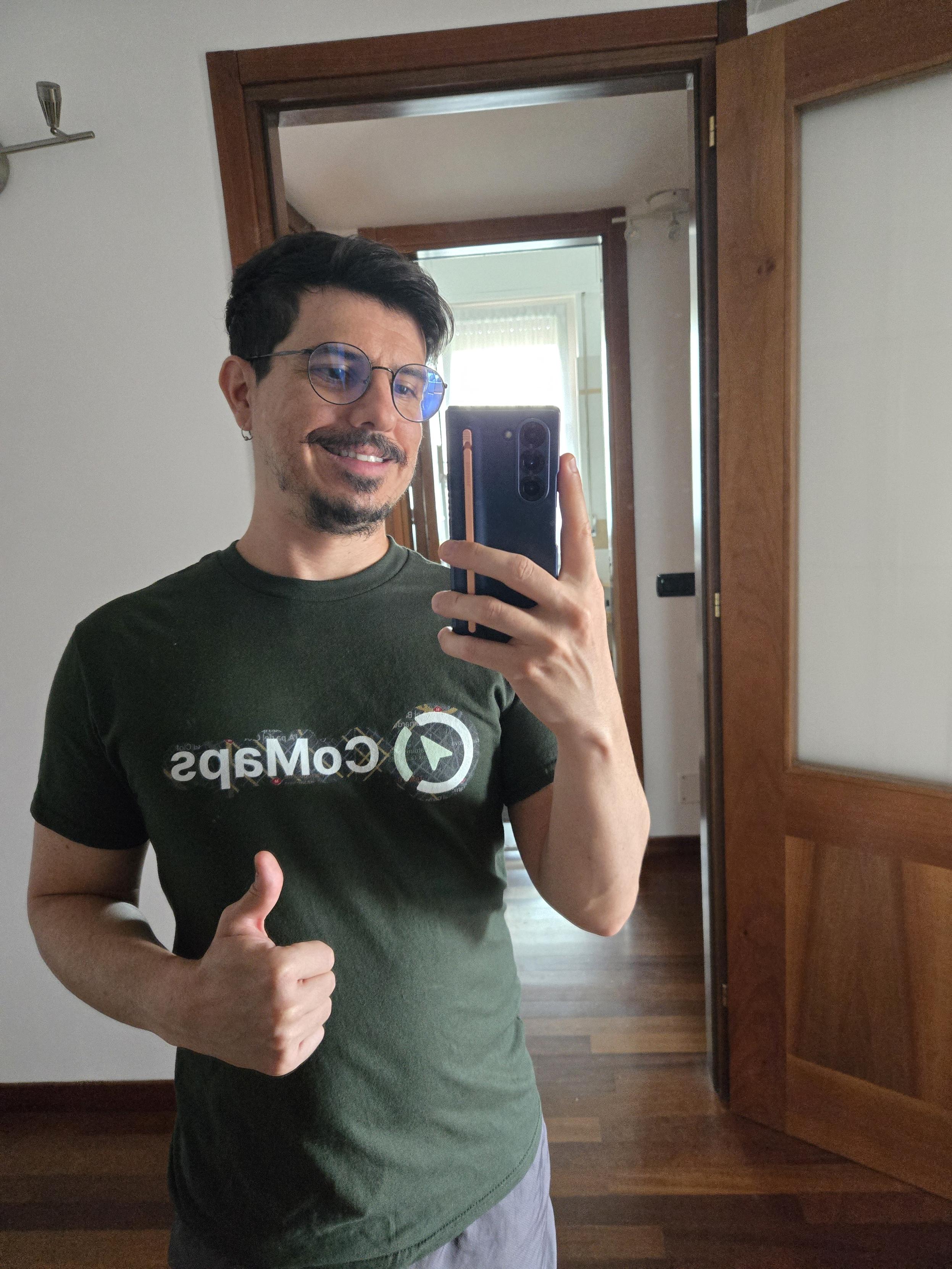 White gay man with a dark green t-shirt printed CoMaps mirrored. His right thumb is up and he is smiling, while looking at his phone to take the picture from his living room mirror.