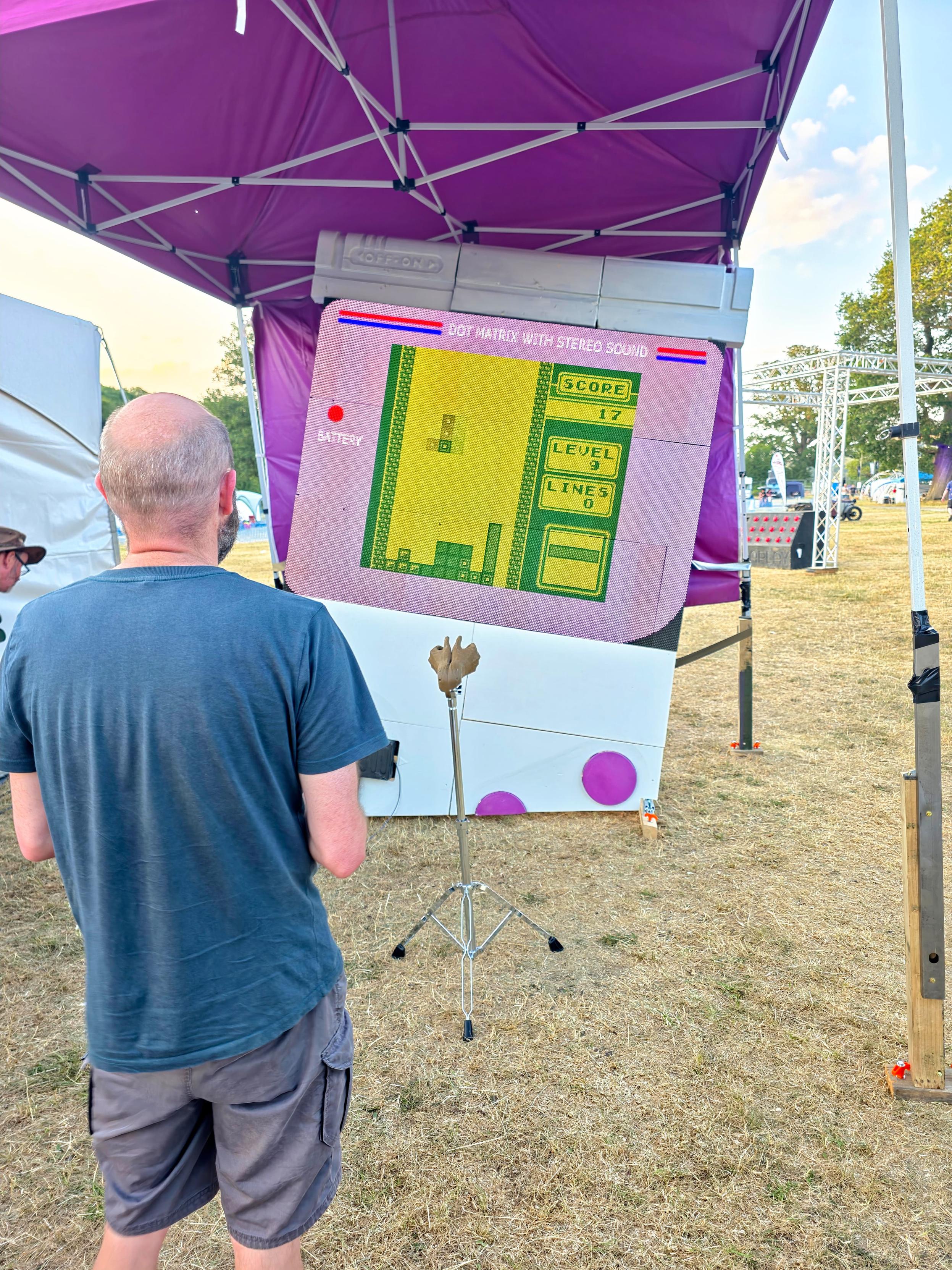 The Hitchin Hackspace Gameboy Monumental is on site at #EMFCamp at last. We didn't quite get it ready in time for the previous event. The picture shows a massive Gameboy which appears to be partially buried, at a tilt to the right, in to the ground. One of the two round buttons on the front of the console is "buried". In front of the console is a music stand, the top of which has been replaced by a pair of 3d printed cupped hands which act as a receptacle or holder for the remote controller for the console. The console is 2.5-3m tall on the screen a game of Tetris is in progress. To the left in the foreground Hitchin Hackspace member Rich stands with his back to the camera.  Unseen his hands hold the controller as he plays the game.