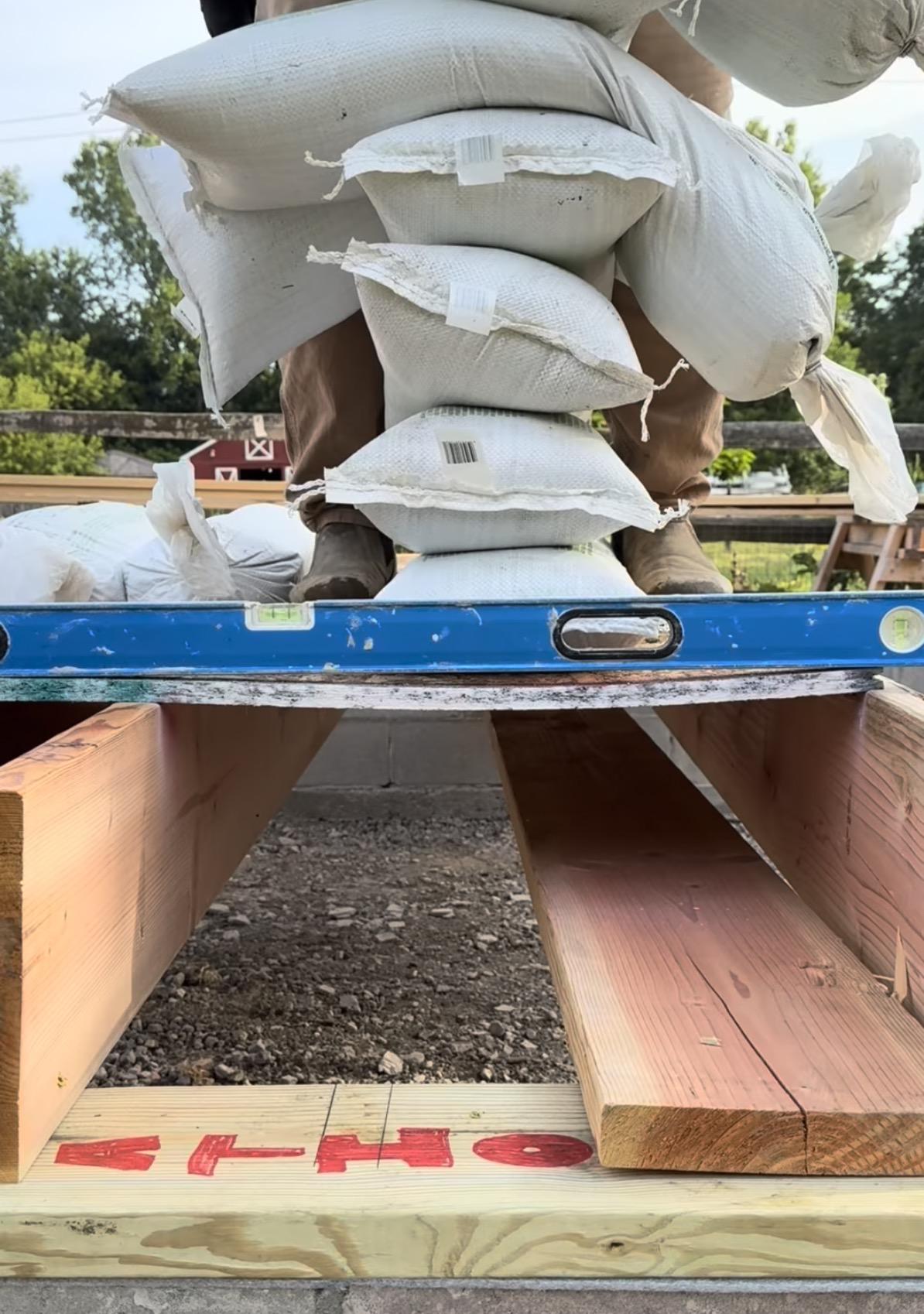 A large pile of sandbags on flooring between joists with a straight edge showing the amount of sag in the middle.