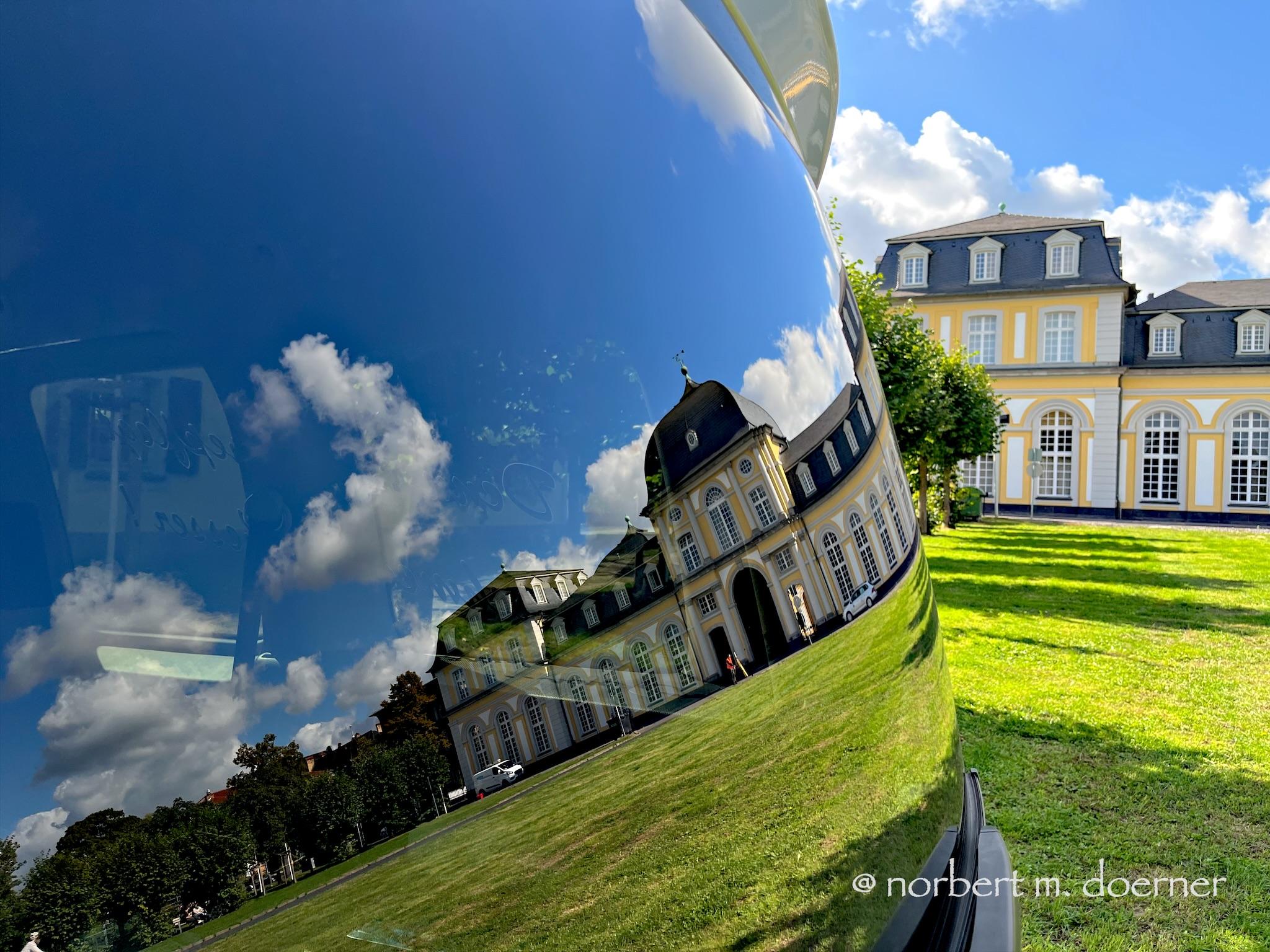 In der gewölbten und fast senkrecht stehenden Heckscheibe eines SUV spiegelt sich ein mit gelben Wänden versehenes Schloss, während man im rechten Drittel des Bildes dieses Gebäude im Original sehen kann, ein wenig entfernt, mit grünem Rasen und darüber blauem Himmel mit einigen wenigen weißen Wölkchen.