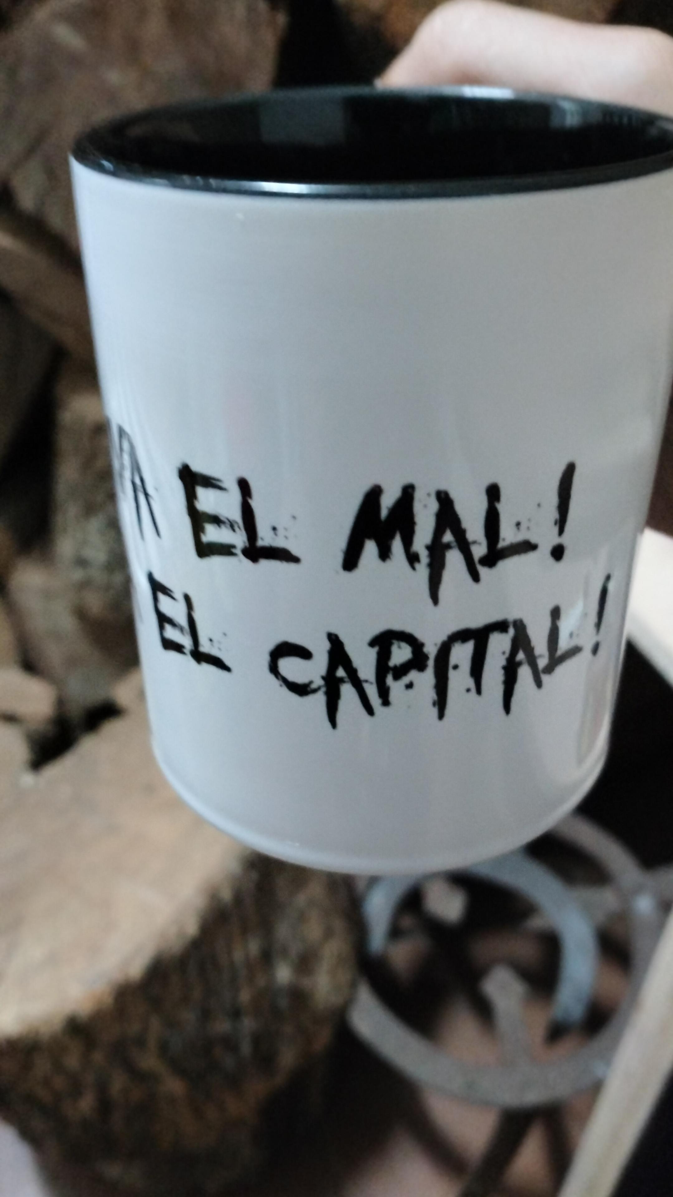 Taza blanca con interior negro. Tiene el lema de la Bruja Avería "¡VIVA EL MAL! ¡VIVA EL CAPITAL!" escrito en negro.
Al fondo se ve, desenfocado, un apilado de leña de encina